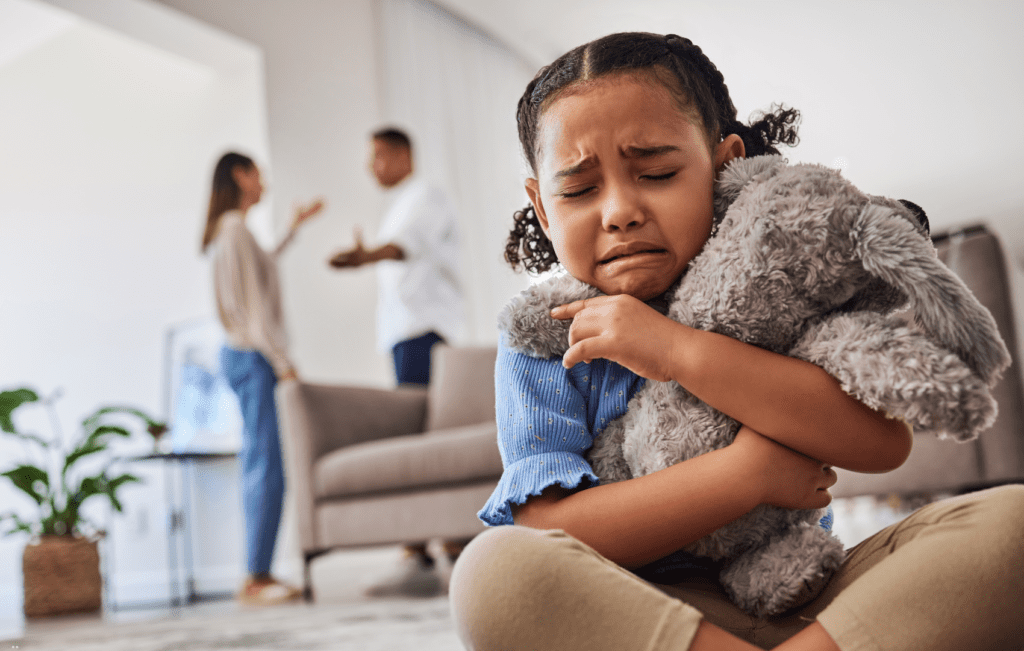 criança abraçando urso de pelúcia e chorando; ao fundo, os pais discutindo, evidenciando a ansiedade infantil