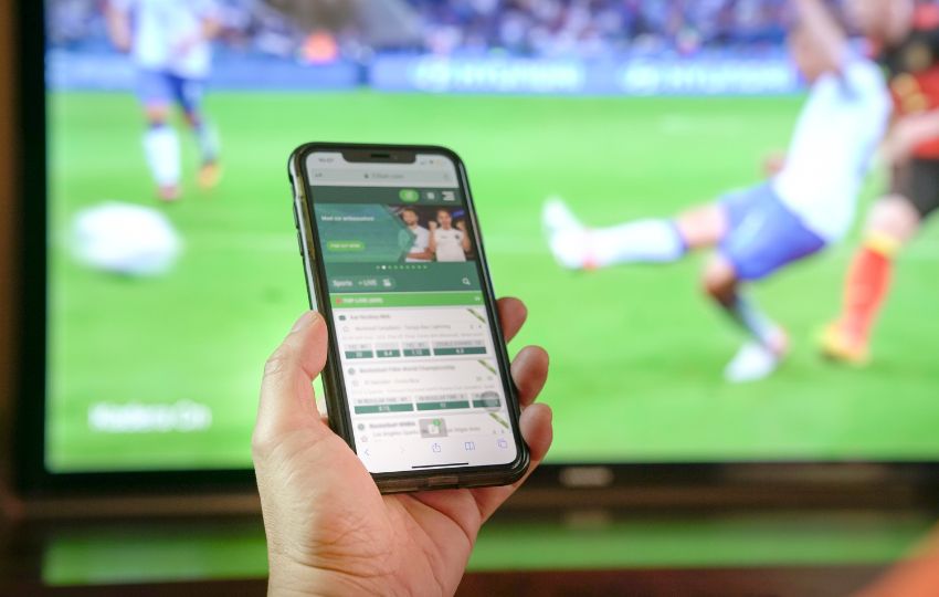 Tela de aplicativo de apostas esportivas no celular, com televisão passando futebol ao fundo, tema associado ao aumento de problemas de saúde mental relacionados às bets.